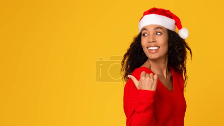 Téléchargez les photos : Joyeux jeune femme noire au chapeau de Père Noël pointant vers l'espace vide, portrait sur fond jaune, idéal pour les offres de Noël, panorama, bannière - en image libre de droit