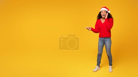 Photo for Surprised black woman wearing Santa hat, energetically pointing at free space, set against vivid yellow backdrop, full length, panorama, banner for Christmas ad - Royalty Free Image