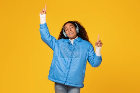 Téléchargez les photos : Jeune femme noire énergique en veste d'hiver dansant tout en écoutant de la musique dans des écouteurs sans fil sur fond jaune, sentant l'ambiance de vacances - en image libre de droit