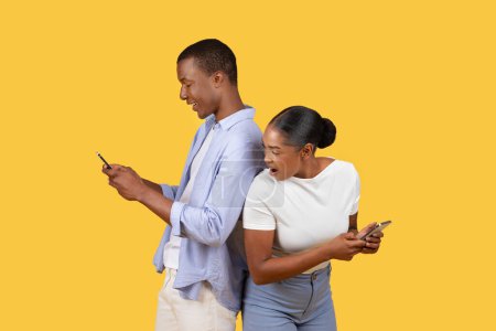 Foto de Curiosa mujer negra se ve sorprendida mientras echa un vistazo al teléfono inteligente mans, ambos de pie espalda con espalda contra el fondo amarillo brillante - Imagen libre de derechos