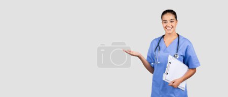Téléchargez les photos : Médecin infirmier millénaire européen positif en uniforme bleu, presse-papiers, espace de copie à portée de main, isolé sur fond de studio gris. Publicité médicale et offre, recommandation de soins de santé - en image libre de droit