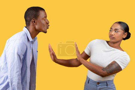 Photo for Black man attempts kiss while the woman holds up her hand in stop sign, showcasing disagreement or boundary setting, yellow background - Royalty Free Image