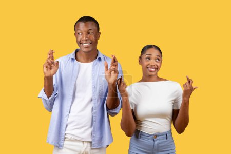 Photo for Optimistic young black man and woman smiling with hopeful expressions, crossing their fingers for good luck against bright yellow background in casual attire - Royalty Free Image