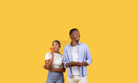 Photo for Pensive woman with credit card and smiling man holding smartphone, both set against bright yellow background, ideal for concepts about shopping and decision-making - Royalty Free Image