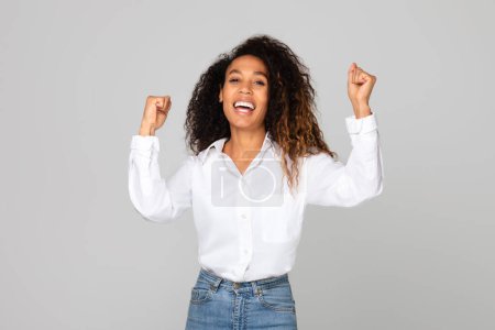 Photo for Cheerful African American Woman Shaking Fists Raising Both Hands, Smiling To Camera, Standing Over Gray Studio Background. Joyful Lady Celebrating Big Luck And Success. Victory Concept - Royalty Free Image