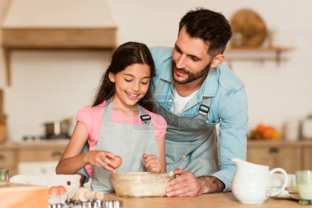 Photo for Caring father teaches his young daughter how to crack an egg into mixing bowl for baking activity in their warm, inviting kitchen, demonstrating shared moment - Royalty Free Image