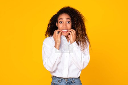 Photo for Worried young black lady expressing fear and anxiety while biting nails isolated on yellow studio background. Expression of tense and overwhelming emotions, response to stress situation - Royalty Free Image