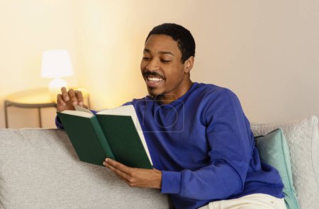Foto de Negro millennial hombre lee libro de bolsillo ahondar en una novela convincente, sentado tranquilamente en su sofá, saboreando su fin de semana en el interior de la casa contemporánea. Bookworm ocio - Imagen libre de derechos