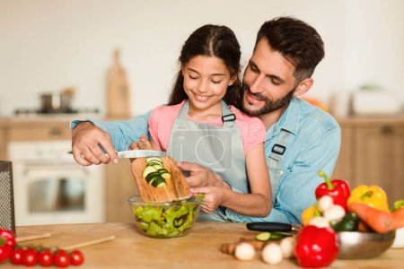 Foto de Padre atento mostrando a su hija cómo cortar pepinos de forma segura para una ensalada en una acogedora cocina casera iluminada por el sol, rodeada de productos frescos. - Imagen libre de derechos