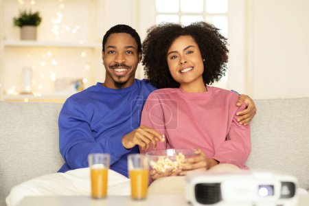 Photo for Black husband and wife enjoy movie and snacks hugging on sofa in living room interior, sitting immersed in a digital domestic cinema theater experience. Weekend quality time - Royalty Free Image