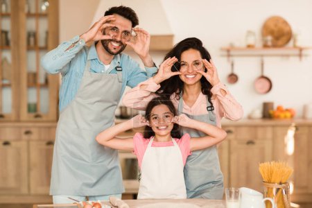 Foto de La familia europea disfruta de un momento lúdico haciendo caras divertidas con cortadores de galletas en una cocina hogareña y bien iluminada, creando recuerdos juntos en casa - Imagen libre de derechos