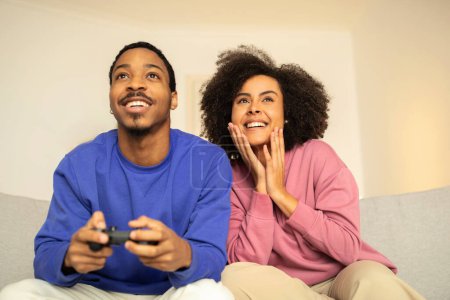 Foto de Emocionados cónyuges afroamericanos absortos en el videojuego digital usando gamepads, compartiendo momentos divertidos, sentados relajados en el interior de su hogar moderno. Vinculación sobre aficiones tecnológicas - Imagen libre de derechos
