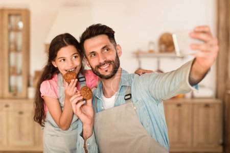 Téléchargez les photos : Père heureux et jeune fille tenant des biscuits faits maison fraîchement cuits aiment prendre selfie ensemble dans un cadre chaleureusement éclairé et confortable cuisine à la maison - en image libre de droit