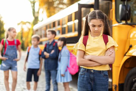Photo for Unhappy young girl standing by herself next to yellow school bus, arms crossed and scowling while other kids laughing and talking behind her, female kid feeling left out and experiencing loneliness - Royalty Free Image