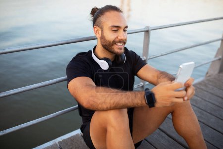 Photo for Sport application. Happy athlete man with headphones texting and scrolling on smartphone, resting from workout while sitting at pier near sea outdoor, exercising on weekend morning - Royalty Free Image