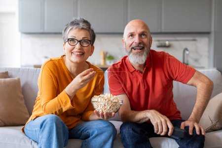 Téléchargez les photos : Portrait d'un couple marié heureux regardant la télévision et mangeant du maïs soufflé à la maison, souriant mari et femme âgés se relaxant sur un canapé confortable dans le salon, bénéficiant d'un film comique - en image libre de droit