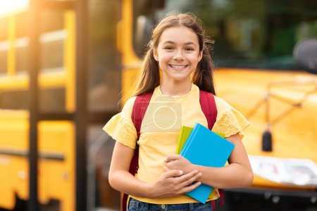 Téléchargez les photos : Retour à l'école. Joyeux sourire fille préadolescente debout près de l'autobus scolaire avec sac à dos et cahiers dans les mains, enfant féminin joyeux posant à l'extérieur tout en allant à des cours, élève enfant appréciant étude - en image libre de droit