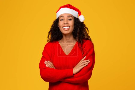Foto de La radiante joven negra con sombrero de Santa Claus posa con confianza con los brazos cruzados, radiante ante en la cámara contra el fondo amarillo brillante, celebrando las vacaciones de Navidad - Imagen libre de derechos