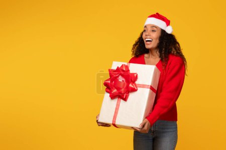 Téléchargez les photos : Jeune femme noire rayonnante dans le chapeau de Père Noël, tenant fièrement grand cadeau de Noël enveloppé, ensemble sur fond jaune vif, espace libre - en image libre de droit