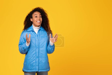 Photo for Surprised young black lady in winter jacket gazes at free space on yellow background, her expression suggesting an incredible holiday offer - Royalty Free Image