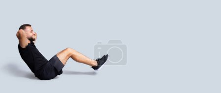 Photo for Smiling handsome strong young caucasian man with beard in sportswear make exercises for abs, isolated on gray studio background, profile, panorama. Body care, sport workout at home, gym - Royalty Free Image