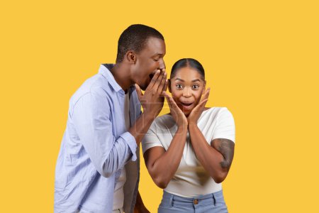 Photo for Black man whispers into womans ear, her expression one of excited shock, against vibrant yellow background - Royalty Free Image