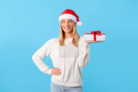 Photo for Cheerful caucasian woman in Santa hat holding out wrapped Christmas present with smile, wearing white sweater, against bright blue background - Royalty Free Image