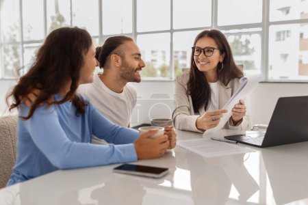 Photo for Happy Homebuyers. Positive Young European Spouses Having Consultation In Real Estate Agency With Middle Aged Woman, Sitting At Desk In Office, Discussing Papers - Royalty Free Image