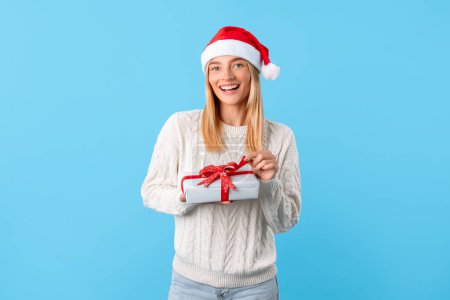 Photo for Delighted woman in cozy sweater and Santa Claus hat holding Christmas present with red ribbon, beaming with joy against soft blue background - Royalty Free Image