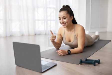 Téléchargez les photos : Une jeune femme saine aux cheveux foncés en tenue de gym pratique le pilates sur son tapis à la maison, regarde l'écran d'ordinateur portable et montre le pouce en l'air, remercie son entraîneur de fitness - en image libre de droit