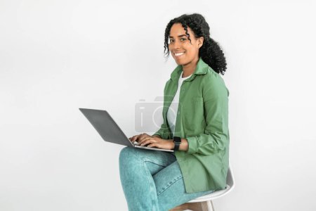 Téléchargez les photos : Jeune femme afro-américaine s'engage dans un travail indépendant, naviguant sur son ordinateur portable tout en étant assis contre un mur de studio blanc, sourire à l'appareil photo jouissant d'un mode de vie numérique - en image libre de droit