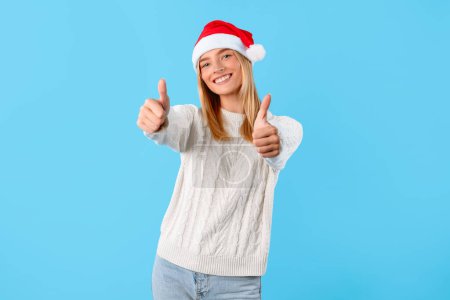 Téléchargez les photos : Jeune femme souriante en chapeau de Noël et pull blanc donne double pouces vers le haut, signalant l'approbation ou le succès, sur fond bleu - en image libre de droit