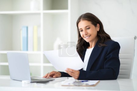 Photo for Positive attractive mature businesswoman wearing formal outfit working at modern office, sitting at desk, typing on laptop, reading documents, checking marketing report, copy space - Royalty Free Image