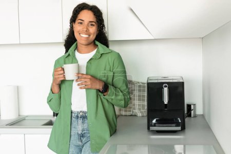Téléchargez les photos : Heureuse jeune femme afro-américaine se tient dans sa cuisine près de la cafetière, avec une tasse de boisson chaude savoureuse dans les mains, mettant en valeur grand début de journée, saveurs moments du matin - en image libre de droit