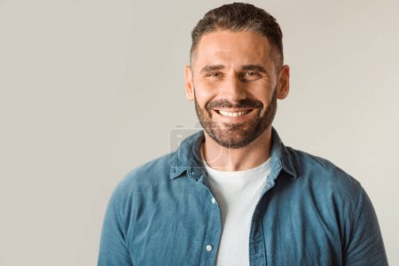 Foto de Hombre de mediana edad en camisas de mezclilla casuales con sonrisa dentada, posando contra la pared gris del estudio, exudando éxito y confianza, mirando a la cámara con expresión amigable. Espacio libre - Imagen libre de derechos