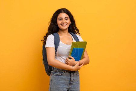 Foto de Concepto de Educación. Mujer india sonriente positiva estudiante sosteniendo cuadernos mirando a la cámara, espacio de copia para la publicidad. Mujer con mochila, fondo amarillo estudio - Imagen libre de derechos