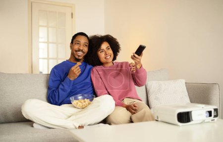 Foto de Sonrientes cónyuges afroamericanos viendo películas en la televisión, comiendo palomitas de maíz mientras cambian de canal en el interior de la sala de estar doméstica, sentados en un sofá juntos y relajados. Concepto romántico de ocio - Imagen libre de derechos