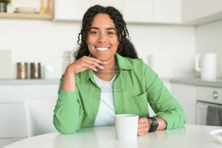 Téléchargez les photos : Portrait de dame noire heureuse dégustant une tasse de café chaud dans la cuisine intérieure de la maison, souriant à la caméra tout en étant assis à table. Femme millénaire excitée ayant pause avec la boisson préférée le week-end - en image libre de droit