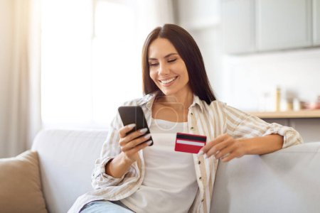 Photo for Happy Young Woman Shopping Online With Smartphone And Credit Card While Relaxing On Sofa At Home, Smiling Female Using Mobile App For Internet Purchases While Resting In Living Room, Copy Space - Royalty Free Image