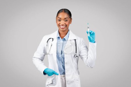 Foto de Mujer negra milenaria positiva en bata blanca, guantes protectores usar jeringa, vacunación de recomendación, aislado en estudio de fondo gris. Medicina e inyección, atención médica - Imagen libre de derechos