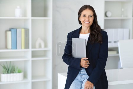 Photo for Cheerful mature woman manager with laptop in her hand standing next desk at office, wearing smart casual outfit. Middle aged corporate businesswoman posing at coworking space, copy space - Royalty Free Image