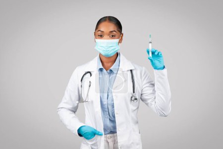 Foto de Sonriente milenaria doctora negra en bata blanca, guantes protectores, jeringa para mascarillas, aislada en un estudio de fondo gris. Recomendación de vacunación, medicina, atención médica en la clínica - Imagen libre de derechos