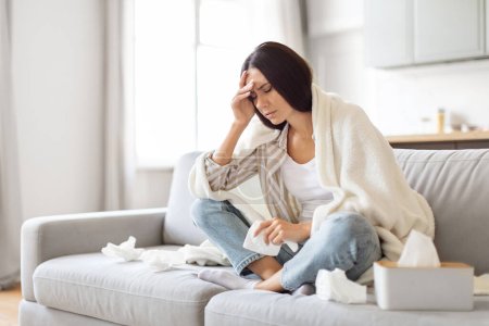 Photo for Seasonal Flu Concept. Sick Young Woman Covered In Blanket Sitting On Couch At Home, Ill Millennial Lady Suffering Headache And Runny Nose, Feeling Unwell, Holding Napkin And Touching Head, Copy Space - Royalty Free Image