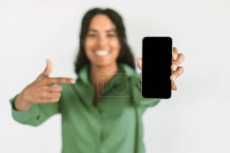Téléchargez les photos : Femme noire en chemise verte présente écran blanc de téléphone portable, pointant vers l'écran tactile sur fond blanc, démontre la facilité de la technologie. Concentration sélective sur le téléphone. Maquette, collage - en image libre de droit