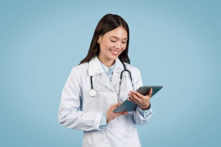 Happy female medical professional in white coat with stethoscope, engaged with digital tablet computer and smiling against clean blue background