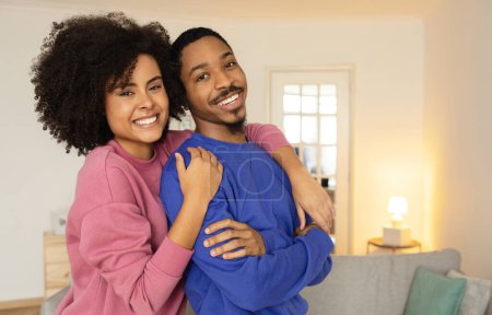 Téléchargez les photos : Les conjoints afro-américains partageant une étreinte chaleureuse et des sourires joyeux, posent pour un portrait chaleureux dans leur salon moderne à la maison, souriant à la caméra. Espace libre pour la publicité - en image libre de droit