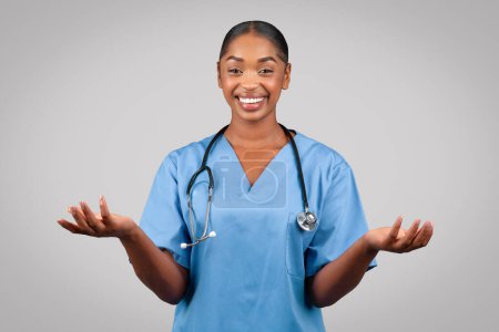 Photo for Positive millennial black doctor surgeon, nurse in blue uniform, spread hands to sides, hold free space on hands, isolated on gray studio background. Medical ad, offer, professional choose - Royalty Free Image
