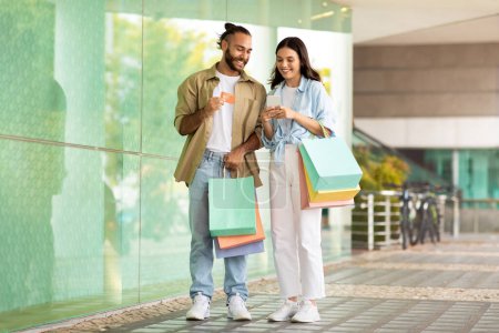 Photo for Cheerful millennial european couple with bags enjoy shopping app on smartphone, credit card in mall, full length. Sale for shopaholics, discount, buy order, cashback and money finance - Royalty Free Image