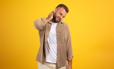 Photo for Unhappy millennial european man with beard in casual massaging neck with hand, suffer from pain, isolated on orange studio background. People emotions, health care problems, spasm - Royalty Free Image