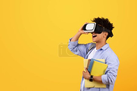 Photo for Stylish black man student engages with virtual reality glasses, standing with notebooks, looking towards empty space on yellow studio background - Royalty Free Image